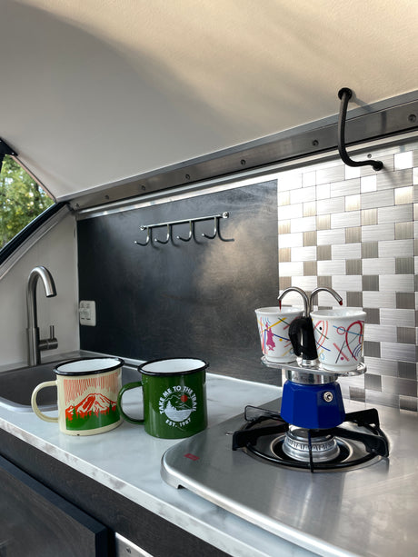Fresh coffee brews in a Moka pot on a stove in a stylish RV rental kitchen, with two enamel mugs waiting.