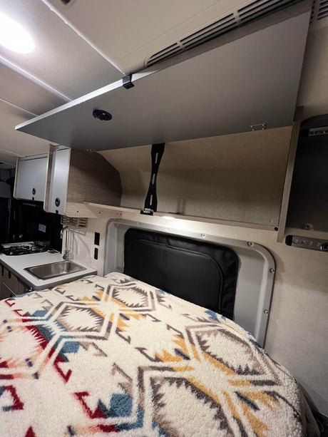 RV rental interior featuring a compact kitchen with sink/stove, comfortable bed with patterned blanket, and open overhead storage.
