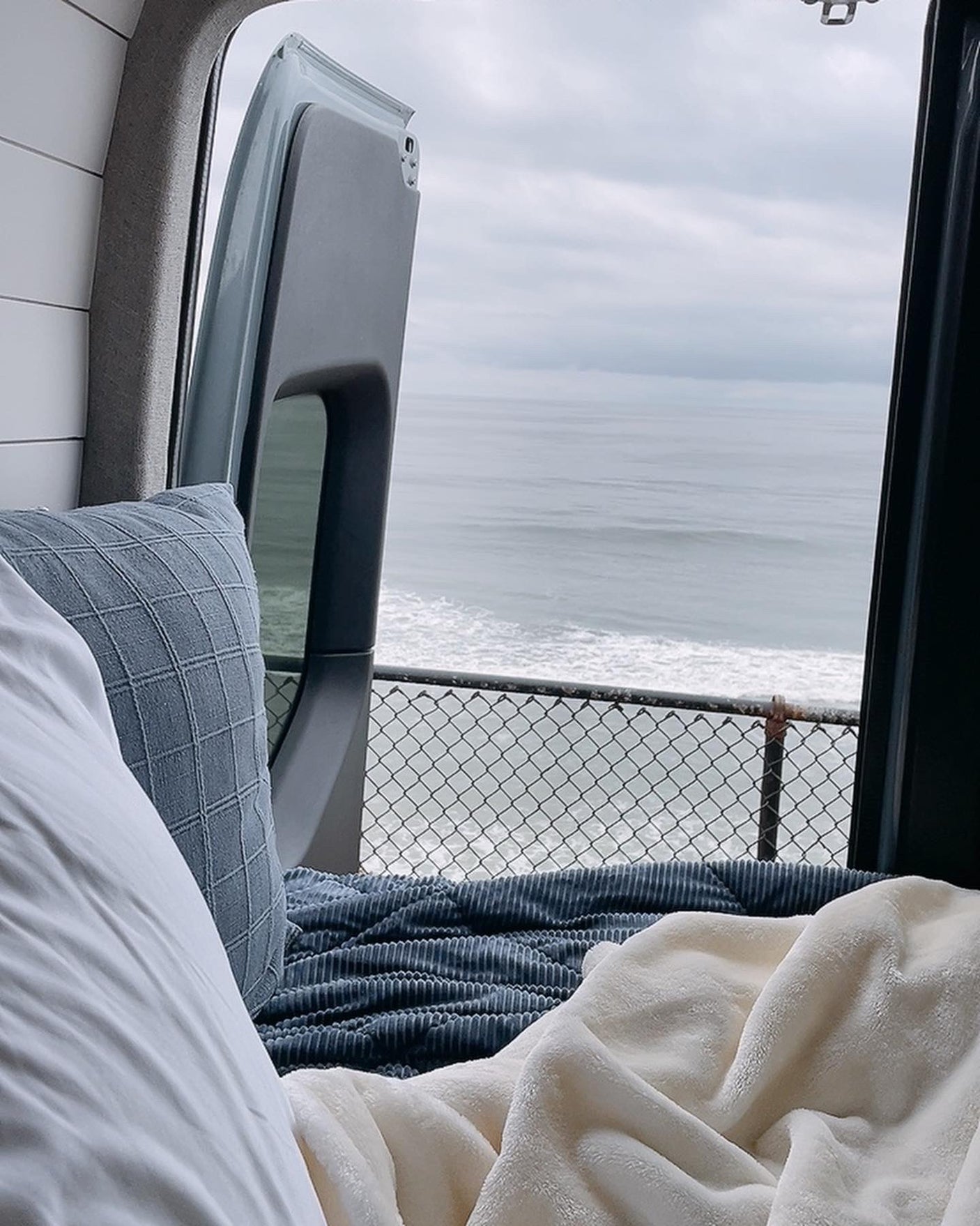 Cozy RV rental bed with a stunning ocean view out the back doors, overlooking crashing waves and a protective fence.