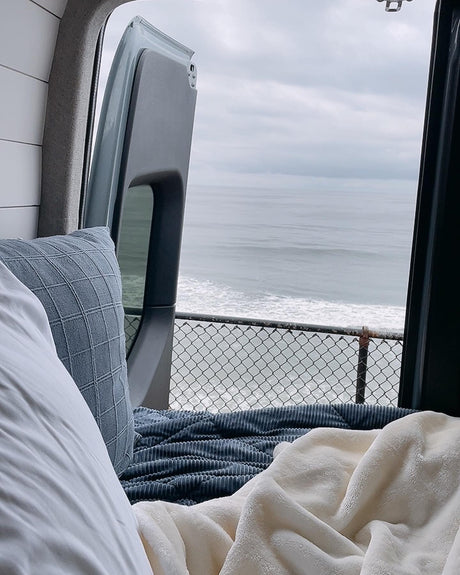 Cozy RV rental bed with a stunning ocean view out the back doors, overlooking crashing waves and a protective fence.