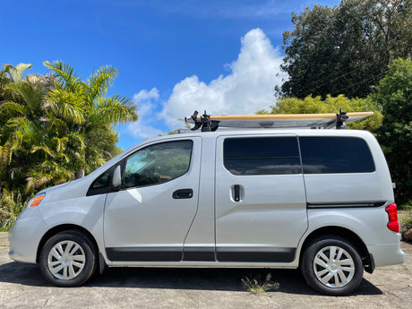 Compact RV rental: A white van with a surfboard on its roof, parked under a blue sky with vibrant palm trees, ready for adventure.