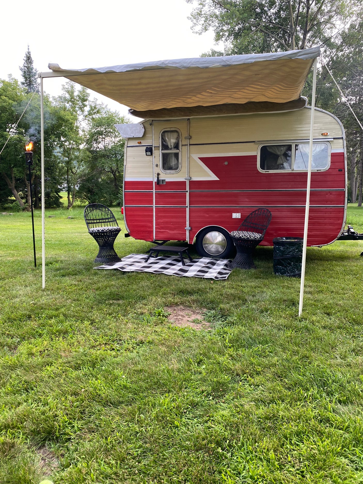 Spacious RV rental setup: vintage camper, extended awning, picnic area with chairs & mat, tiki torch.