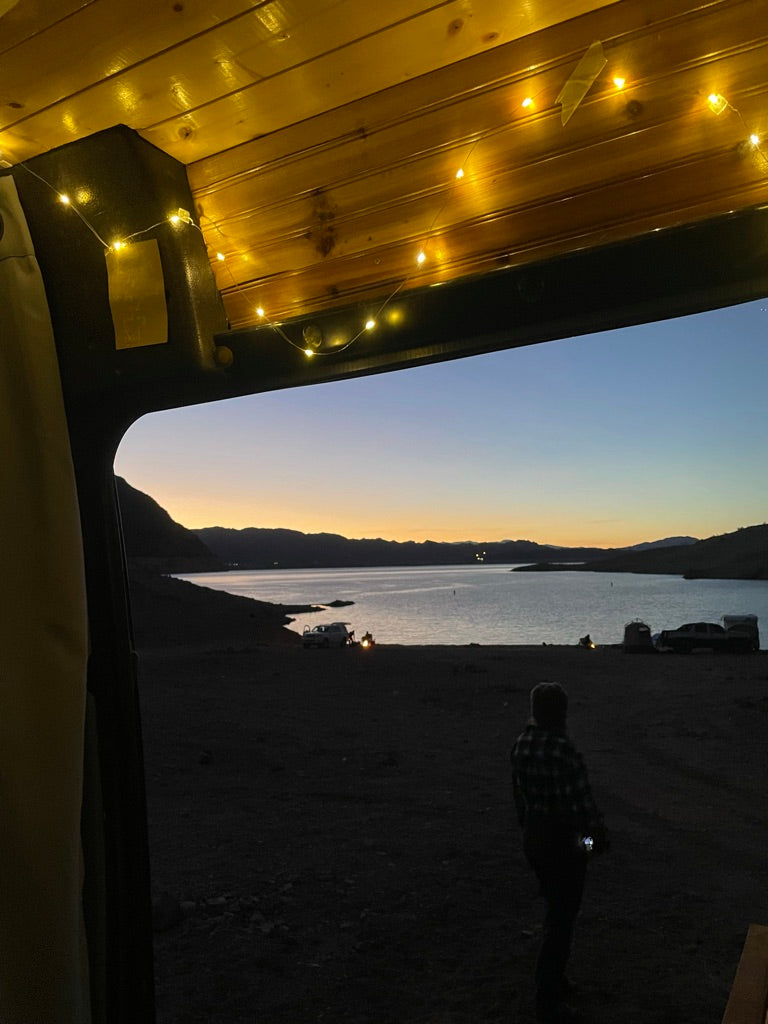 Peaceful lake camping from an RV rental. Dusk view shows mountains, a serene lake, and a person outside, with cozy interior string lights.
