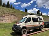 Storyteller Overland van, an adventure RV rental, parked on a grassy hill with tall trees under a bright sky.
