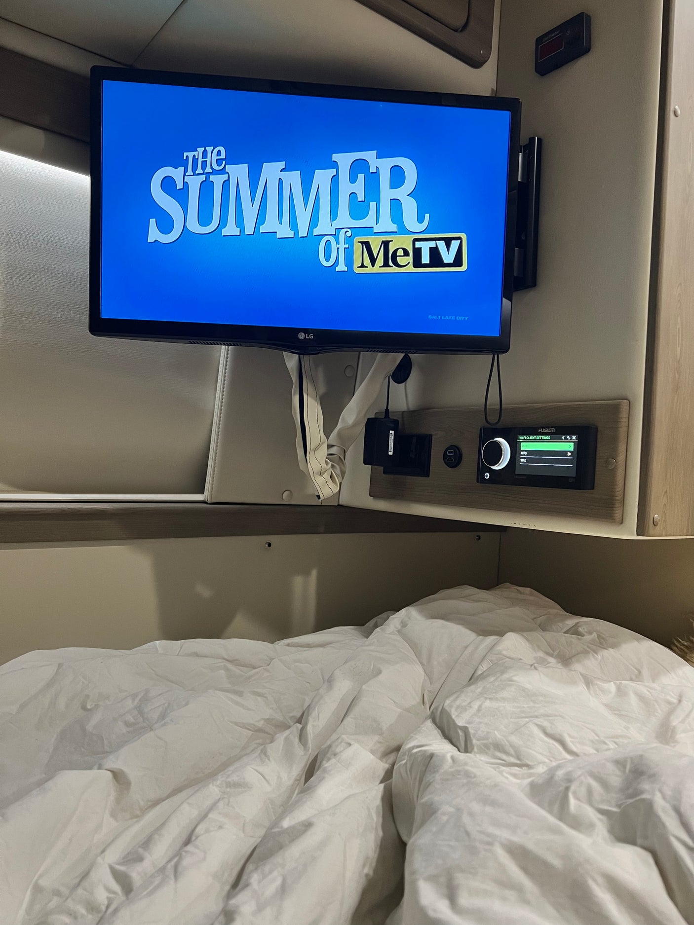 Inside a comfy rv rental, a TV plays MeTV above a bed. Controls for Wi-Fi and power are integrated into the wall.