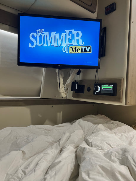 Inside a comfy rv rental, a TV plays MeTV above a bed. Controls for Wi-Fi and power are integrated into the wall.