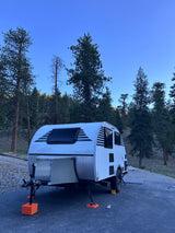 This teardrop RV rental, a compact camper, is set up for a serene camping trip among tall pine trees under a clear sky.