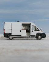 White camper van, door open to reveal interior kitchen, parked on a light, vast plain under blue sky. Perfect for an RV rental adventure.