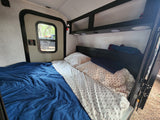 RV rental interior: A made bed with blue and patterned linens, a side door with a window, and overhead storage with lights.