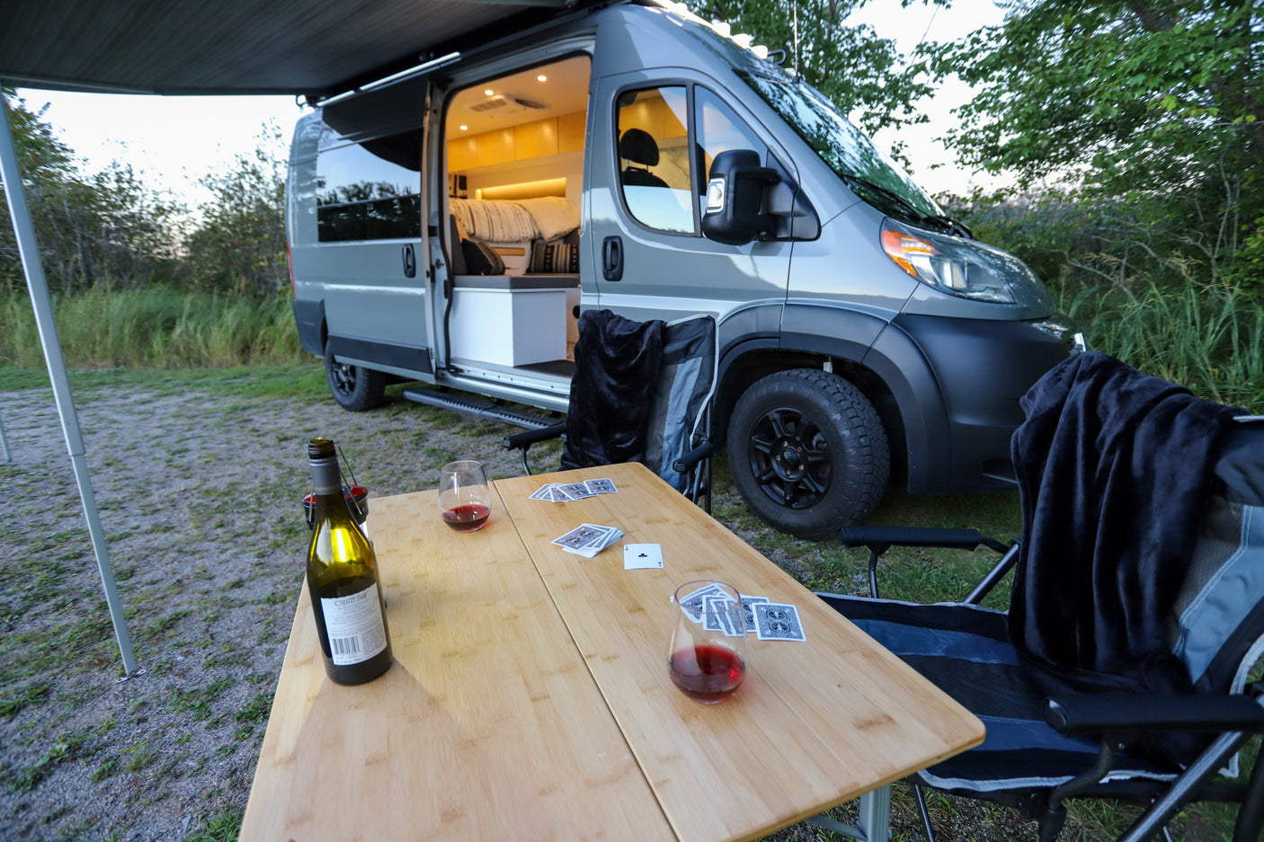 This cozy RV rental scene features a camper van, extended awning, table with wine & cards for a relaxing outdoor evening.