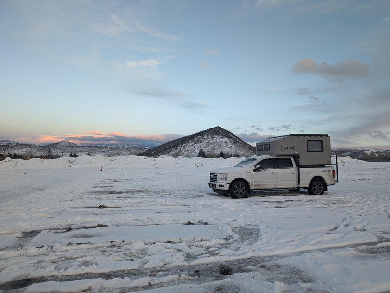 A white Ford pickup with a grey Scout truck camper is parked on a snowy plain with mountains in the background. Consider an RV rental for winter camping. — photo 20