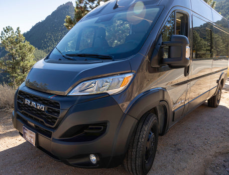 Dark gray Ram ProMaster camper van for RV rental, parked on a dirt road with mountains & trees in the background.