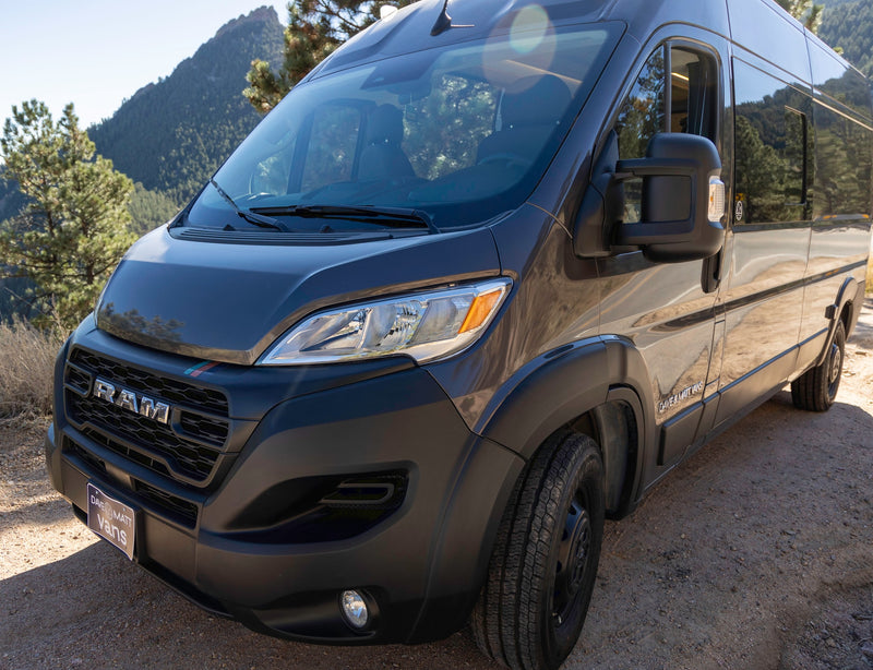 Dark gray Ram ProMaster camper van for RV rental, parked on a dirt road with mountains & trees in the background. — photo 5