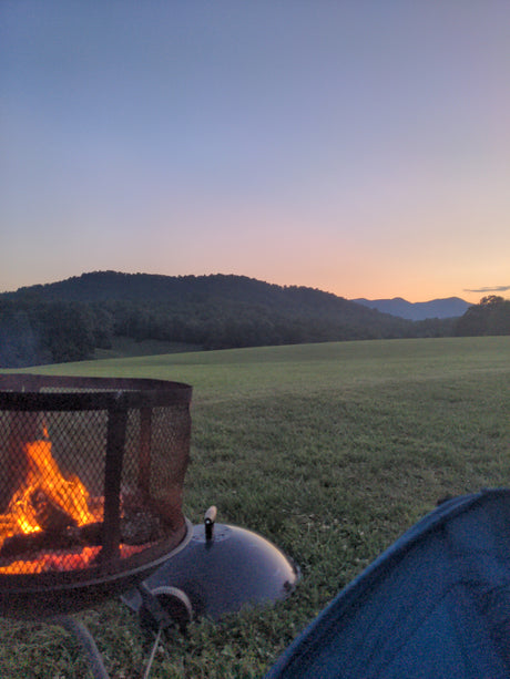 Warm campfire glowing at sunset with mountains & a field. Perfect evening at your RV rental spot, enjoying nature's beauty.