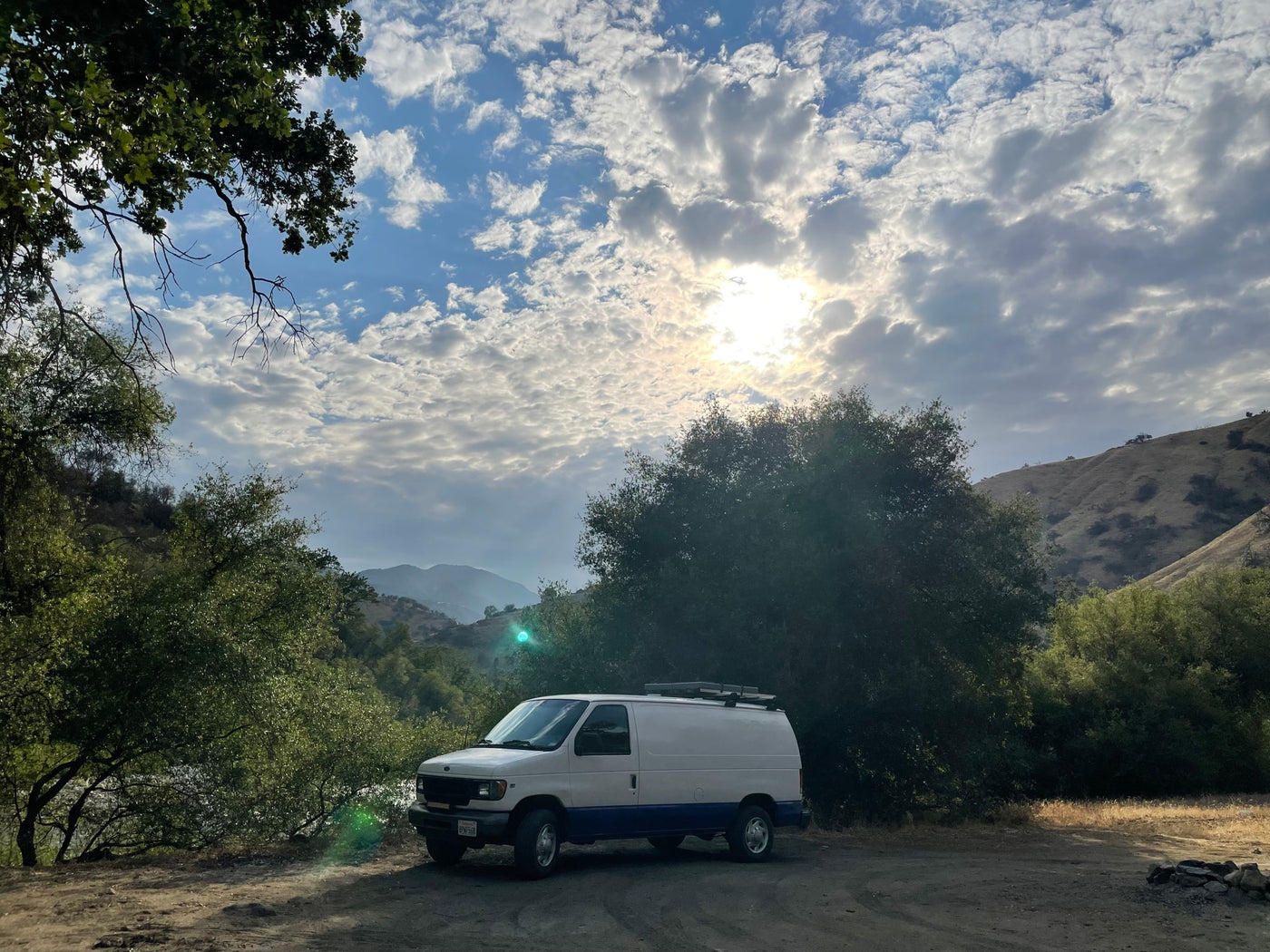 Scenic spot for an RV rental! A white and blue camper van sits in a rustic setting with trees, mountains, and a cloudy, sunlit sky. — photo 8