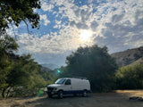 Scenic spot for an RV rental! A white and blue camper van sits in a rustic setting with trees, mountains, and a cloudy, sunlit sky.