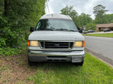 White Ford RV rental parked roadside. Raised roof camper van, headlights on, surrounded by trees and grass.