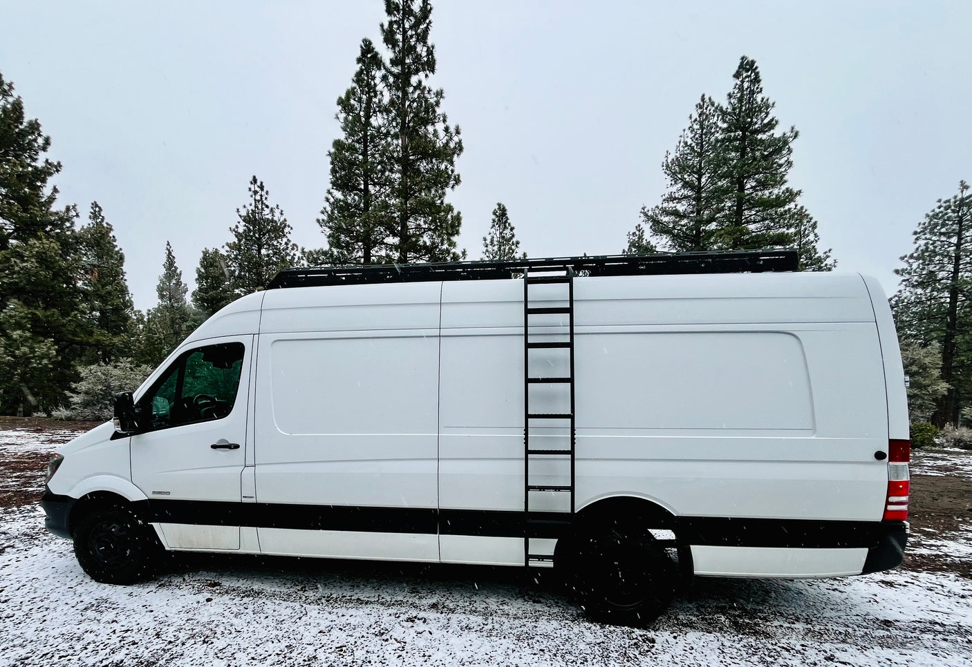 White Sprinter van RV rental with a roof rack and ladder, parked in a snow-covered pine forest. Ideal for winter travel.