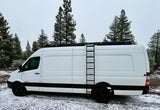 White Sprinter van RV rental with a roof rack and ladder, parked in a snow-covered pine forest. Ideal for winter travel.