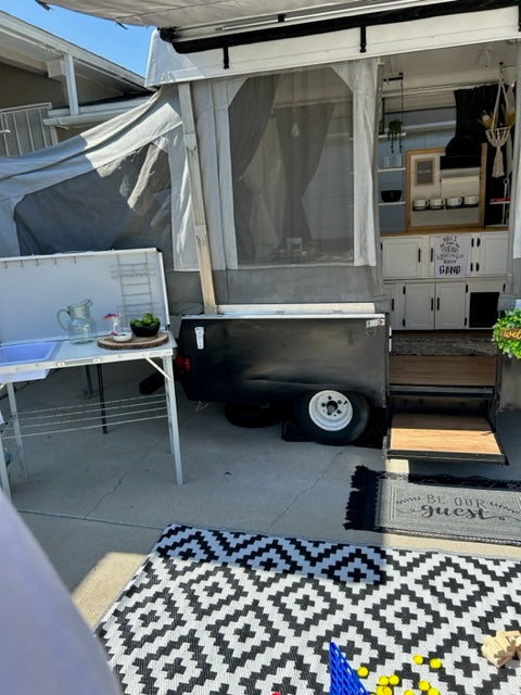 An RV rental, a pop-up camper, fully set up with an outdoor kitchen, patio rug, and interior cabinets visible.