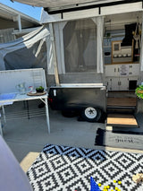 An RV rental, a pop-up camper, fully set up with an outdoor kitchen, patio rug, and interior cabinets visible.