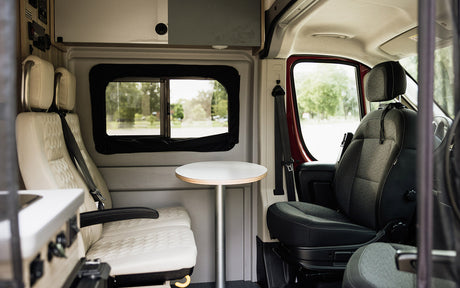 Inside this modern RV rental, see passenger and driver seats, a small table, and a window view. Ideal for travel.