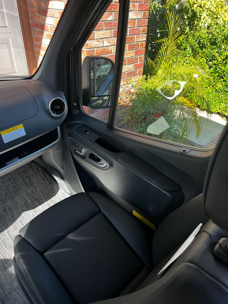 Bright RV rental interior: black passenger seat, dashboard, and door. Sunny window view reveals green plants and a brick house.