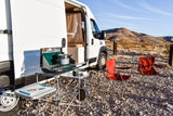 RV rental camp setup: white van, outdoor stove with pot, cutting board, red chairs on rocky ground, desert views.