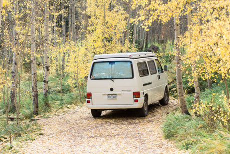 White camper van on a yellow leaf-covered road, framed by tall aspen trees with bright autumn foliage. Ideal RV rental experience.
