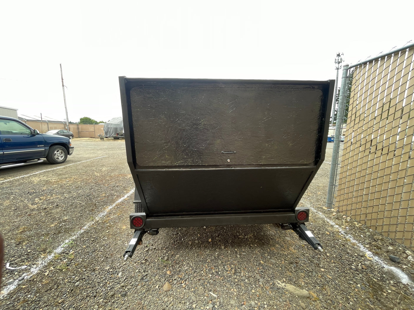 Back of a dark gray utility trailer with red tail lights and stabilizing jacks on a gravel lot near a fence. Suitable for RV rental operations.