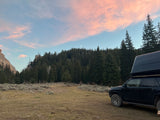 RV rental adventure! Sunset wilderness camping with an SUV & rooftop tent, nestled among mountains and a forest under a crescent moon.