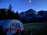 Nighttime view: A teardrop RV rental parked amidst towering mountains and dark pine trees under a bright moon.