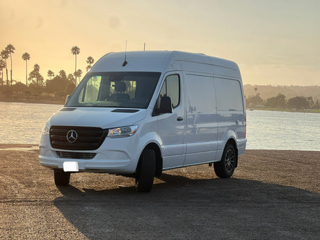 White Mercedes Sprinter van, perfect for your next RV rental adventure, parked by water & palm trees at sunset.
