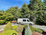 Light gray RV rental van with a rooftop awning, towing an orange raft on a trailer. Surrounded by lush trees and a blue sky.