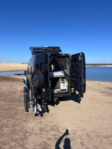 Black camper van RV rental with bike, parked by a scenic lake. Open rear doors reveal custom interior, ready for adventure!