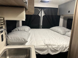 Cozy RV rental interior featuring a comfy bed with white sheets, a small kitchenette with a sink, and overhead storage.