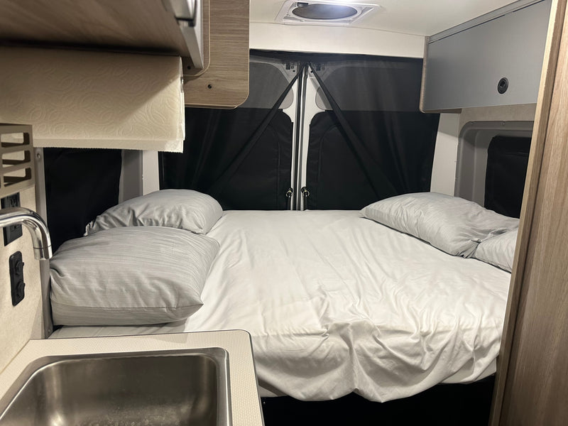 Cozy RV rental interior featuring a comfy bed with white sheets, a small kitchenette with a sink, and overhead storage. — photo 12