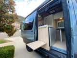Gray RV rental camper van with open side door. Features mobile kitchen, fridge, and fold-out outdoor table. Perfect for adventures!