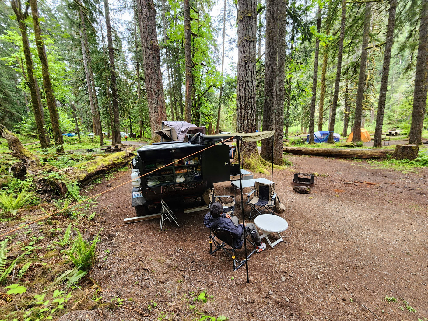 Cozy forest RV rental campsite: black adventure vehicle with open kitchen, rooftop tent, awning, chairs, and a person relaxing.
