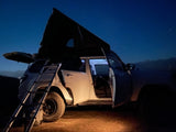 White SUV with rooftop tent & ladder, an ideal RV rental for car camping under a starry night sky. Illuminated interior.