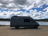 Spacious dark grey camper van on dirt by a lake, perfect for an RV rental adventure under a cloudy sky.
