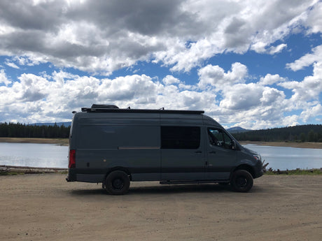 Spacious dark grey camper van on dirt by a lake, perfect for an RV rental adventure under a cloudy sky.