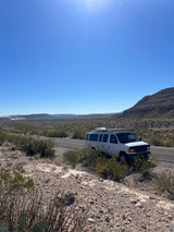 An RV rental van is parked off a desert road, surrounded by dry brush & mountains, under a bright blue sky.