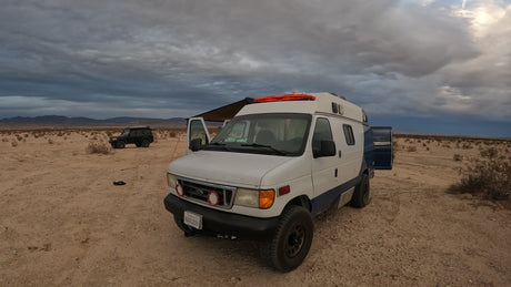 White off-road RV rental van with awning & a second SUV parked in a vast, sandy desert under cloudy skies. Rugged adventures.