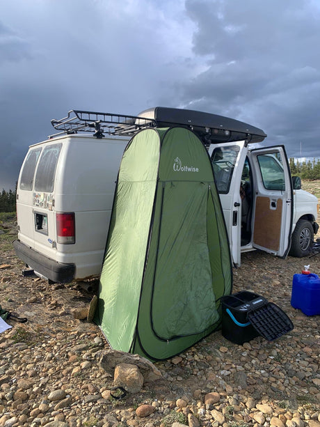An RV rental van parked on rocky terrain with a privacy tent, portable toilet, and roof storage for remote camping.