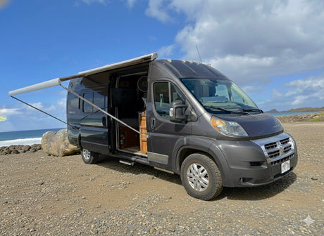 Dark gray RV rental camper van with awning open on a rocky beach by the ocean under a blue sky.