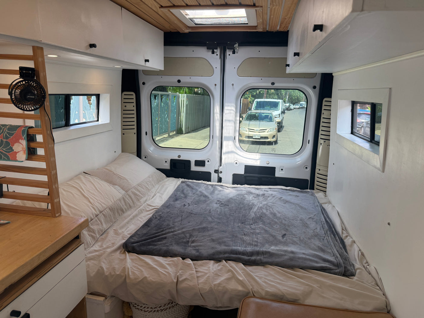 Cozy RV rental interior: a comfy bed, wooden shelves with a fan, and a view of the street through the open rear doors.