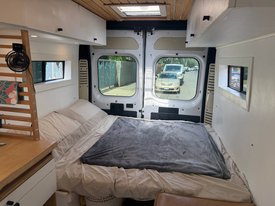 Cozy RV rental interior: a comfy bed, wooden shelves with a fan, and a view of the street through the open rear doors.