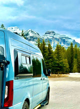 Light blue camper van RV rental parked. Snowy peaks and dense pine trees in the scenic mountain background.