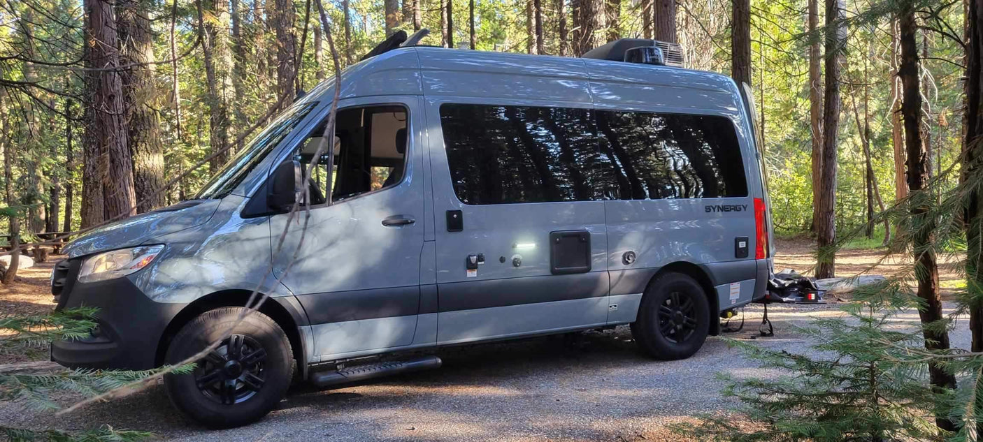 This sleek gray rv rental camper van sits ready for adventure at a sunny forest campsite, surrounded by tall trees.